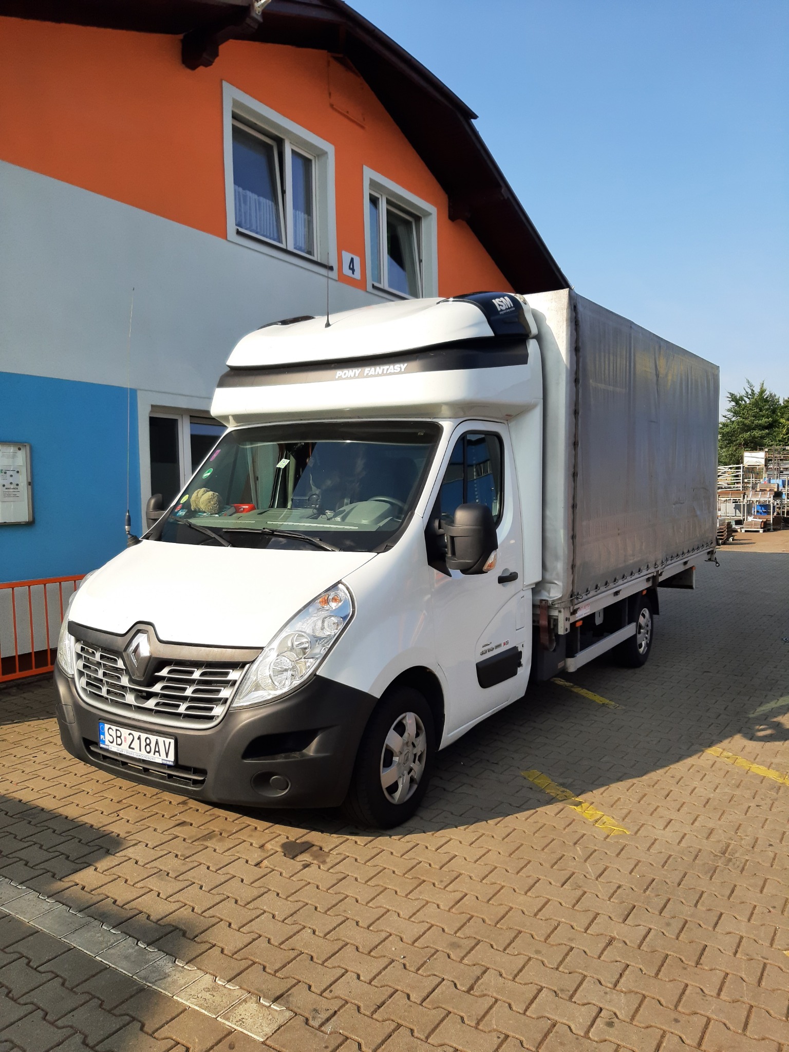 Biały samochód dostawczy Renault Master z plandeką, zaparkowany na kostce brukowej z żółtymi liniami, na tle budynku z pomarańczowo-niebieskimi elementami.