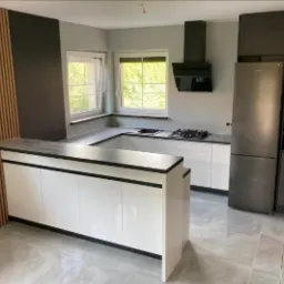 Nowoczesna kuchnia w odcieniach szarości i bieli z drewnianymi panelami ściennymi, wyspą kuchenną z czarnym blatem, oknami z widokiem na zieleń i płytkami podłogowymi imitującymi marmur.