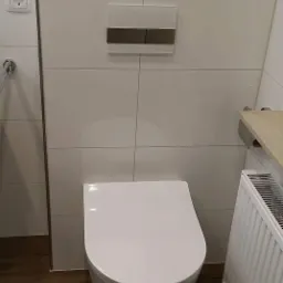 Nowoczesna toaleta podwieszana z białymi płytkami i drewnopodobną podłogą, widoczny grzejnik i przycisk spłuczki w kolorze białym i brązowym.