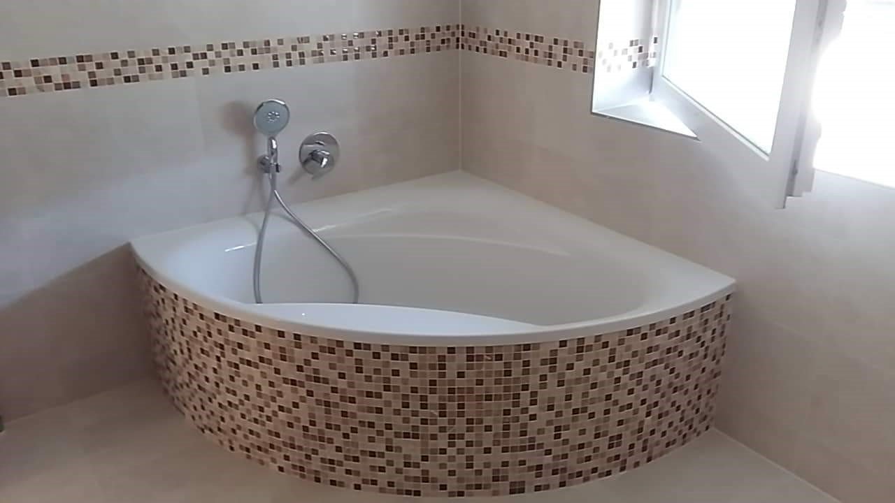 Narożna wanna obudowana mozaiką w odcieniach brązu i beżu, z chromowaną słuchawką prysznicową i baterią na ścianie, uzupełniona pasem mozaiki pod sufitem.