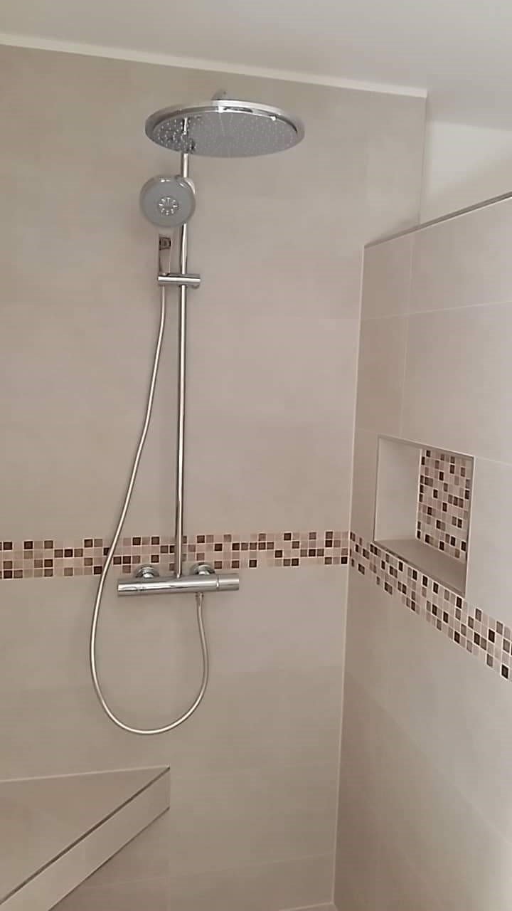 Nowoczesny prysznic z deszczownicą i baterią termostatyczną, wykończony jasnymi płytkami z dekoracyjnym pasem mozaiki i wnęką.