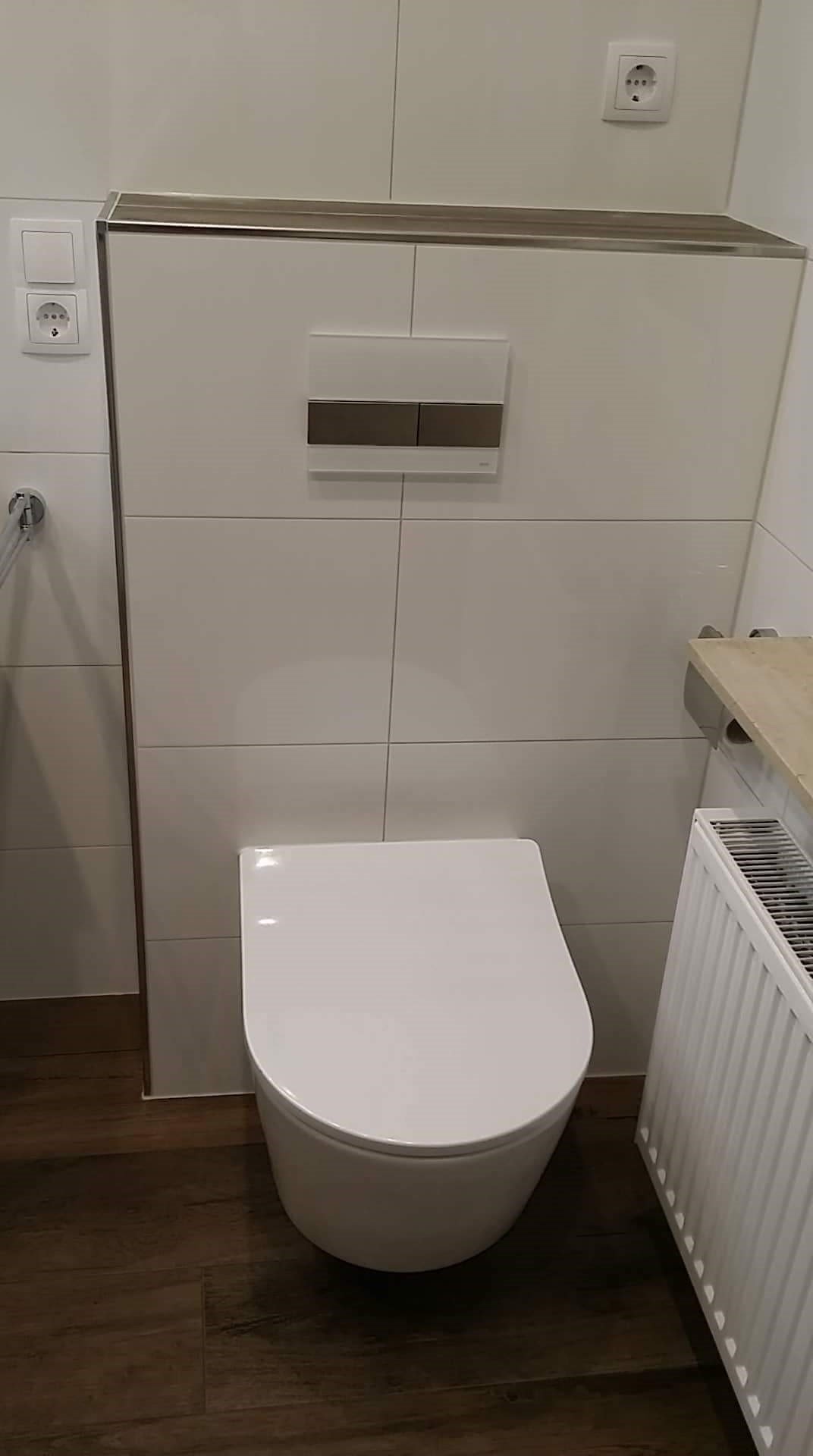 Nowoczesna toaleta podwieszana z białymi płytkami i drewnopodobną podłogą, widoczny grzejnik i przycisk spłuczki w kolorze białym i brązowym.