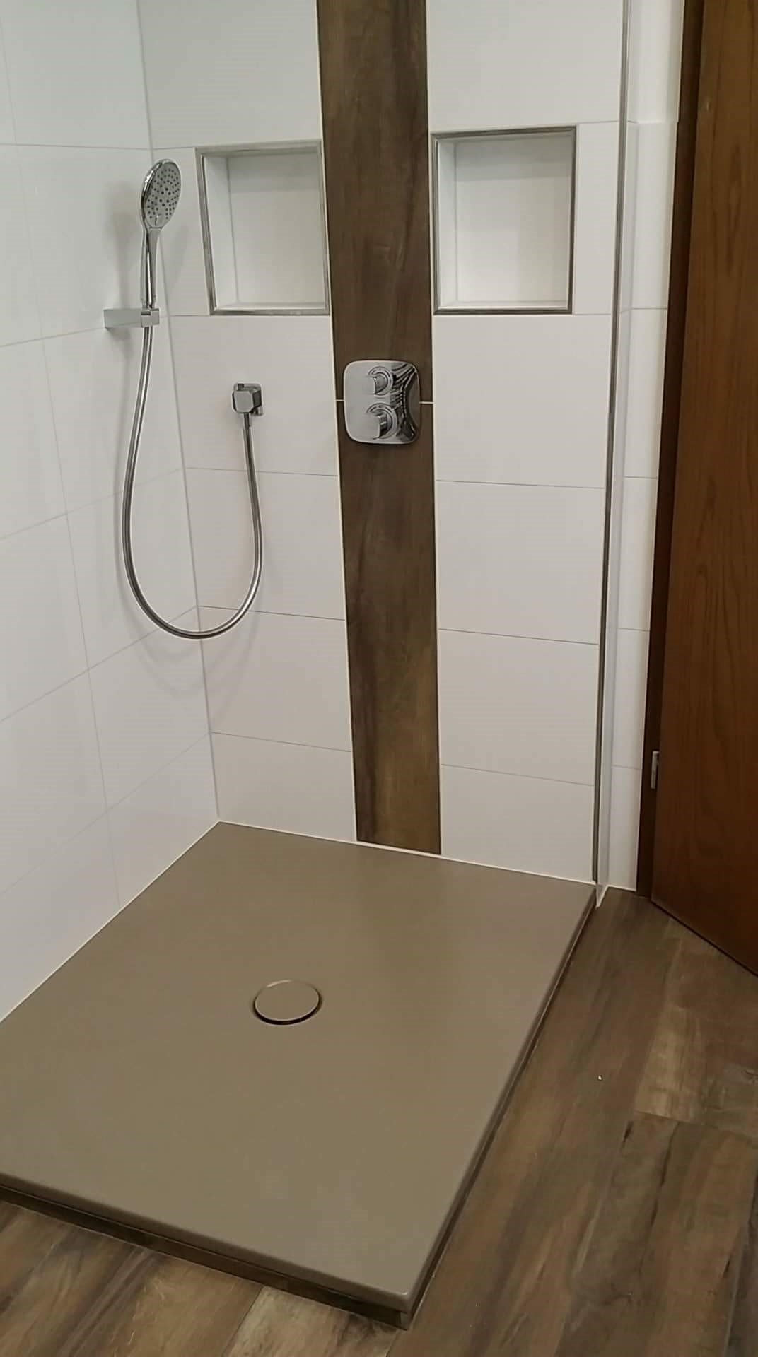 Nowoczesna kabina prysznicowa z odpływem liniowym, wykończona białymi płytkami z wnękami i panelem imitującym drewno, bateria prysznicowa z deszczownicą.