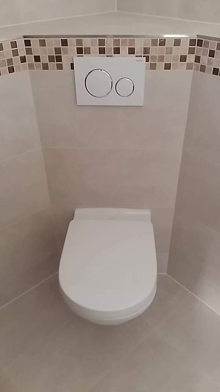 Kompaktowa toaleta podwieszana z dwoma przyciskami spłukującymi, zamontowana w narożniku wykończonym płytkami w odcieniach beżu i brązu, z dekoracyjnym pasem mozaiki nad przyciskami.