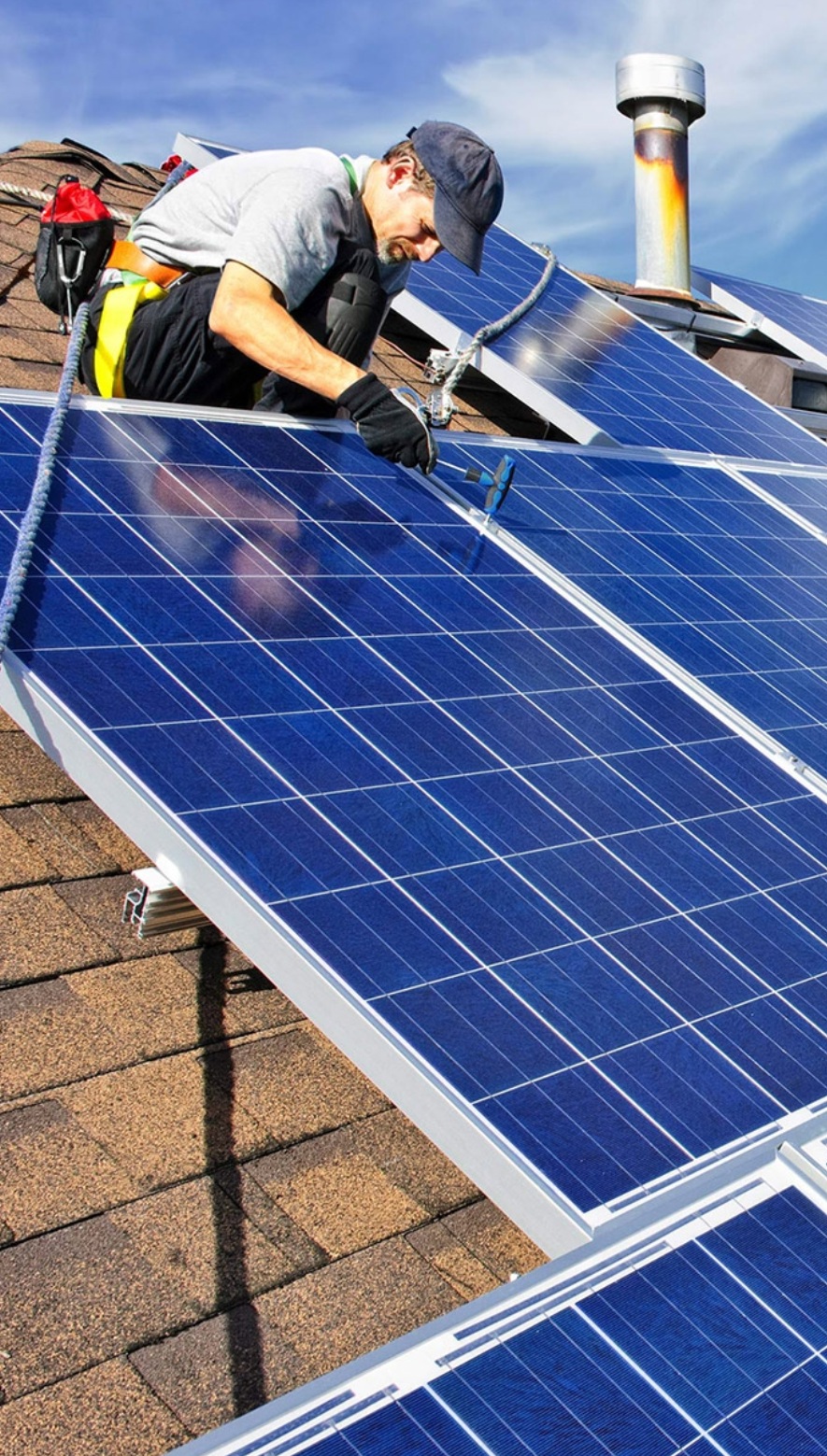 Pracownik w uprzęży zabezpieczającej montuje panele słoneczne na brązowym dachu domu, używając niebieskiego narzędzia do wyrównywania paneli; w tle komin i błękitne niebo.