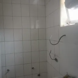 Świeżo wykafelkowana ściana łazienki z widocznymi instalacjami elektrycznymi i hydraulicznymi przed montażem armatury, z fragmentem umywalki w rogu kadru i pojemnikiem z narzędziami na niej