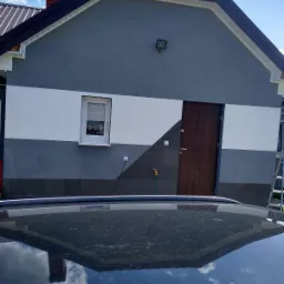 Dom z ocieploną i pomalowaną elewacją w odcieniach szarości i bieli, widoczna drewniana stolarka drzwiowa i okienna oraz odbicie domu w dachu samochodu.