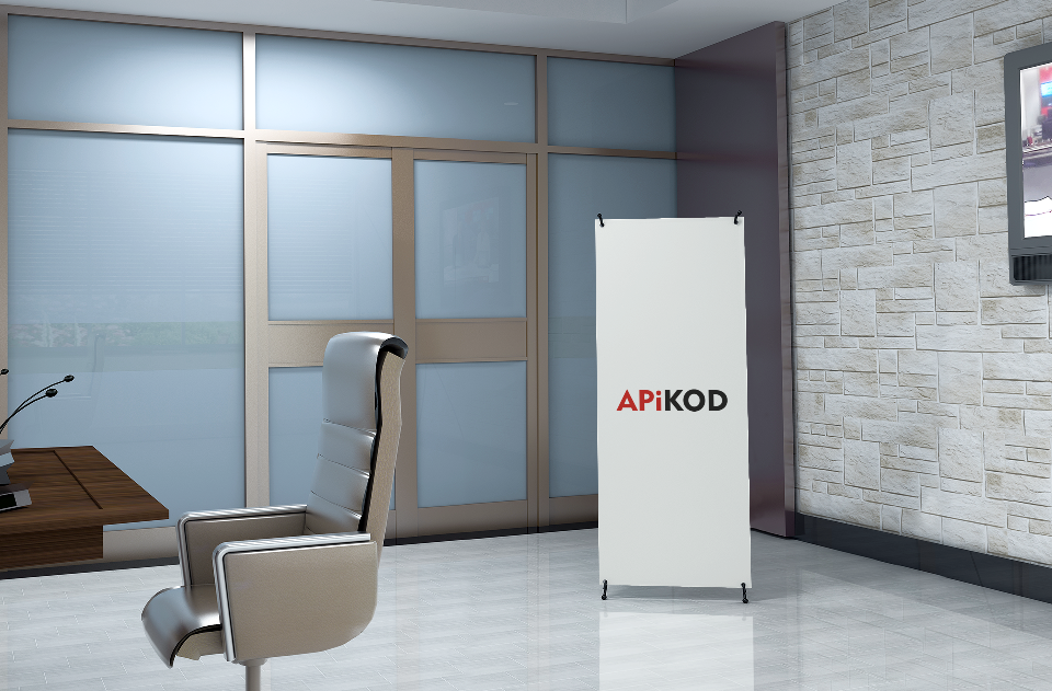 Wnętrze biura z ergonomicznym fotelem, biurkiem i roll-upem z logo ApiKOD na tle okna i ściany z cegły.