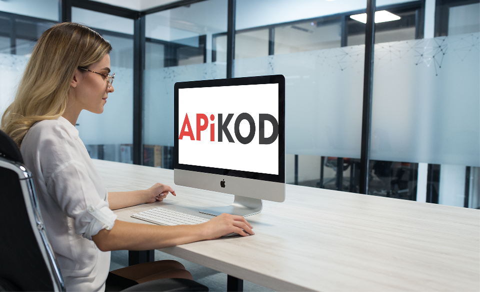 Kobieta w okularach siedzi przed komputerem iMac, na ekranie widoczne logo 'APiKOD' w kolorach czerwonym i czarnym, biuro z przeszklonymi ścianami w tle.