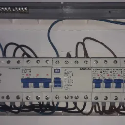 Rozdzielnica elektryczna z zainstalowanymi bezpiecznikami Schrack, widoczne kable w kolorach niebieskim i brązowym, listwy zaciskowe na górze i po prawej stronie.