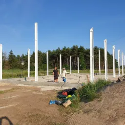 Fundament z białymi słupami konstrukcyjnymi w trakcie budowy domu murowanego, dwóch pracowników w tle, narzędzia i materiały budowlane na pierwszym planie.