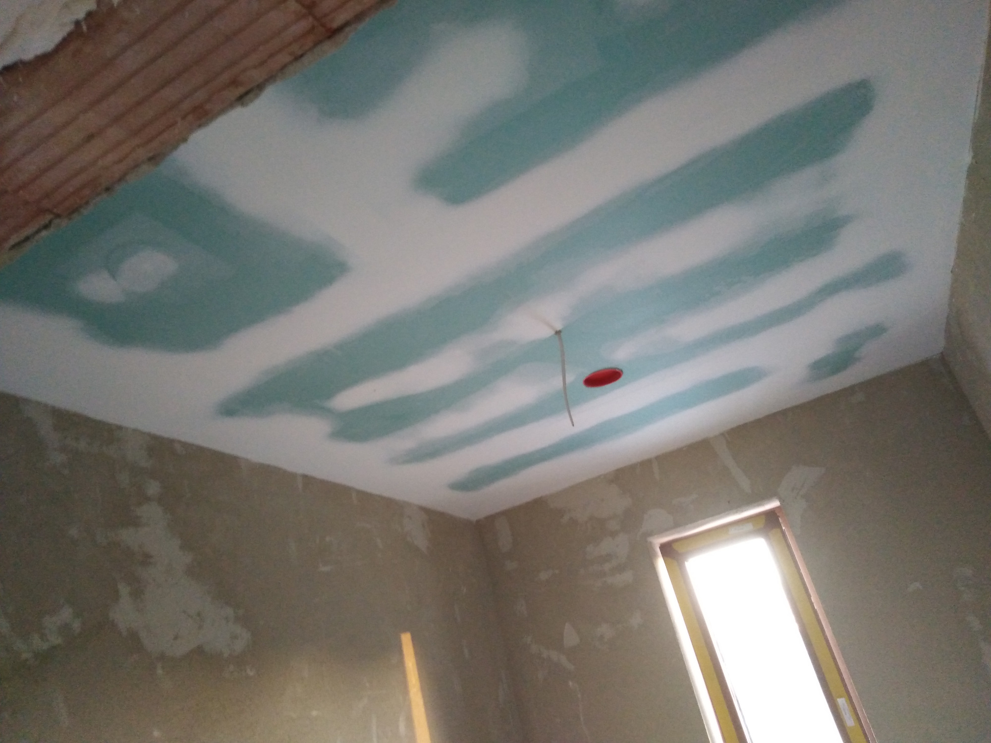 Nierównomiernie nałożona warstwa szpachli na suficie, widoczne ślady po szpachlowaniu, surowy stan ścian w pomieszczeniu z oknem.