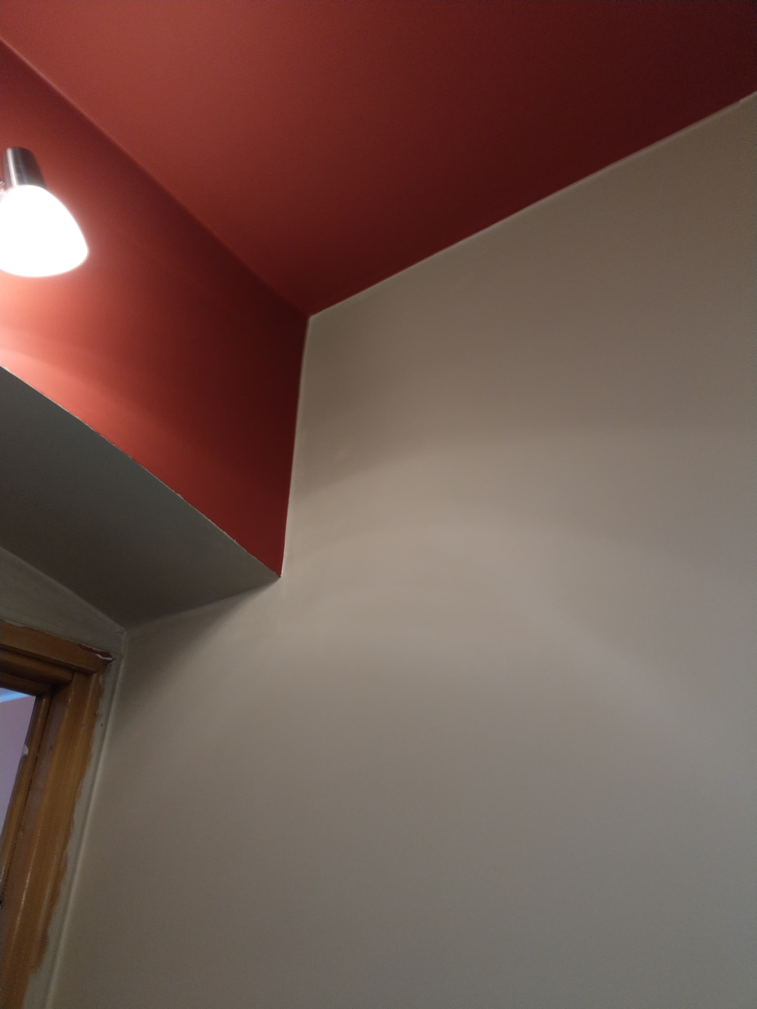 Pomarańczowy sufit i beżowa ściana w rogu pokoju, widoczna lampa sufitowa i fragment ościeżnicy drzwi.