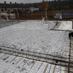 Montaż ogrzewania podłogowego: białe rury ułożone w pętle na izolacji z siatką, wzmocnione stalową konstrukcją zbrojeniową, widok z góry na placu budowy.