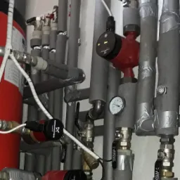 Szare rury instalacji grzewczej z zaworami i manometrem, izolowane termicznie, z widocznymi czerwonymi pompami obiegowymi Grundfos i zbiornikiem wyrównawczym.