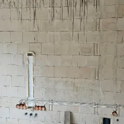 Surowa ściana z betonu komórkowego z widocznymi przewodami elektrycznymi w puszkach, pionowy kanał wypełniony pianką montażową, oraz fragment instalacji hydraulicznej z niebieskimi rurami w otworze...