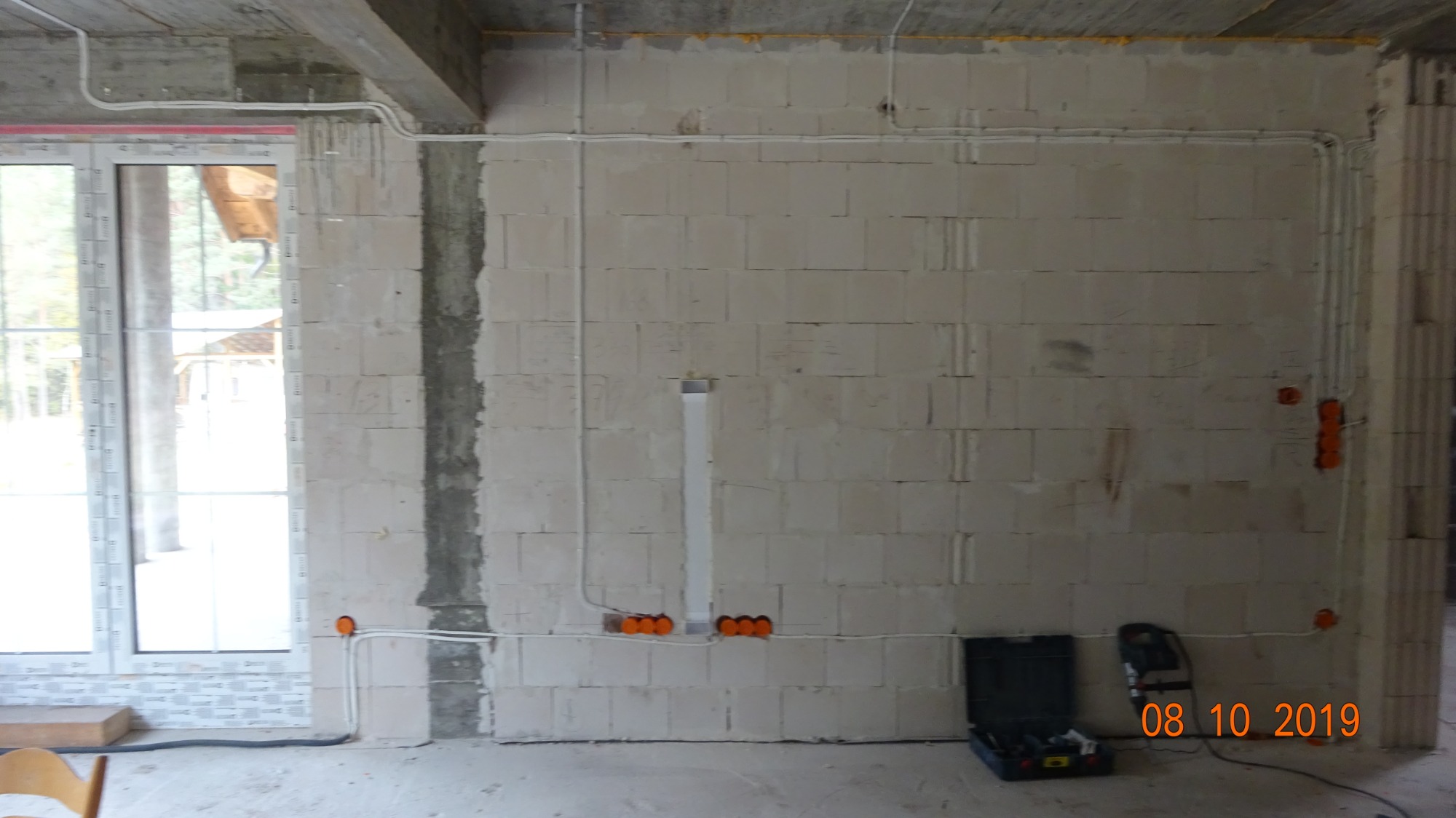 Surowa ściana z betonu komórkowego z widocznymi przewodami elektrycznymi w peszlach, puszkami instalacyjnymi i bruzdami pod tynk, z narzędziami w skrzynce leżącymi na podłodze.