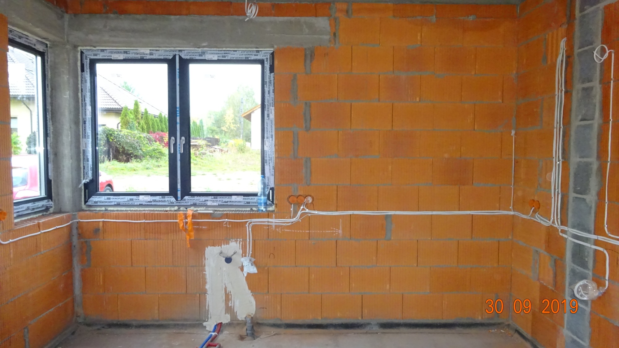 Wnętrze w trakcie prac elektrycznych: ściany z cegły dziurawki, okna PCV, rozprowadzone przewody elektryczne w peszlach, przygotowane punkty pod gniazdka i włączniki, ślady po szpachlowaniu przy...