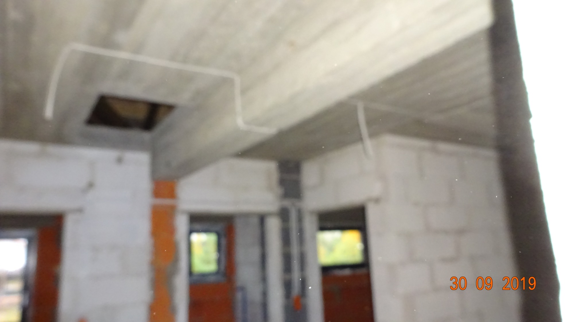 Surowy stan wnętrza budynku w trakcie budowy z widocznymi przewodami elektrycznymi pod sufitem i otworami okiennymi.