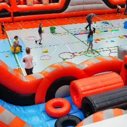 Wielki, pomarańczowo-szary dmuchany tor przeszkód w hali sportowej, dzieci pokonują różne przeszkody, w tle widoczny duży dmuchany zamek.