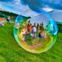 Dzieci i dorośli bawią się na trawie, tworząc gigantyczne bańki mydlane, widoczne w odbiciu dużej bańki na pierwszym planie, na tle białego namiotu i pochmurnego nieba.