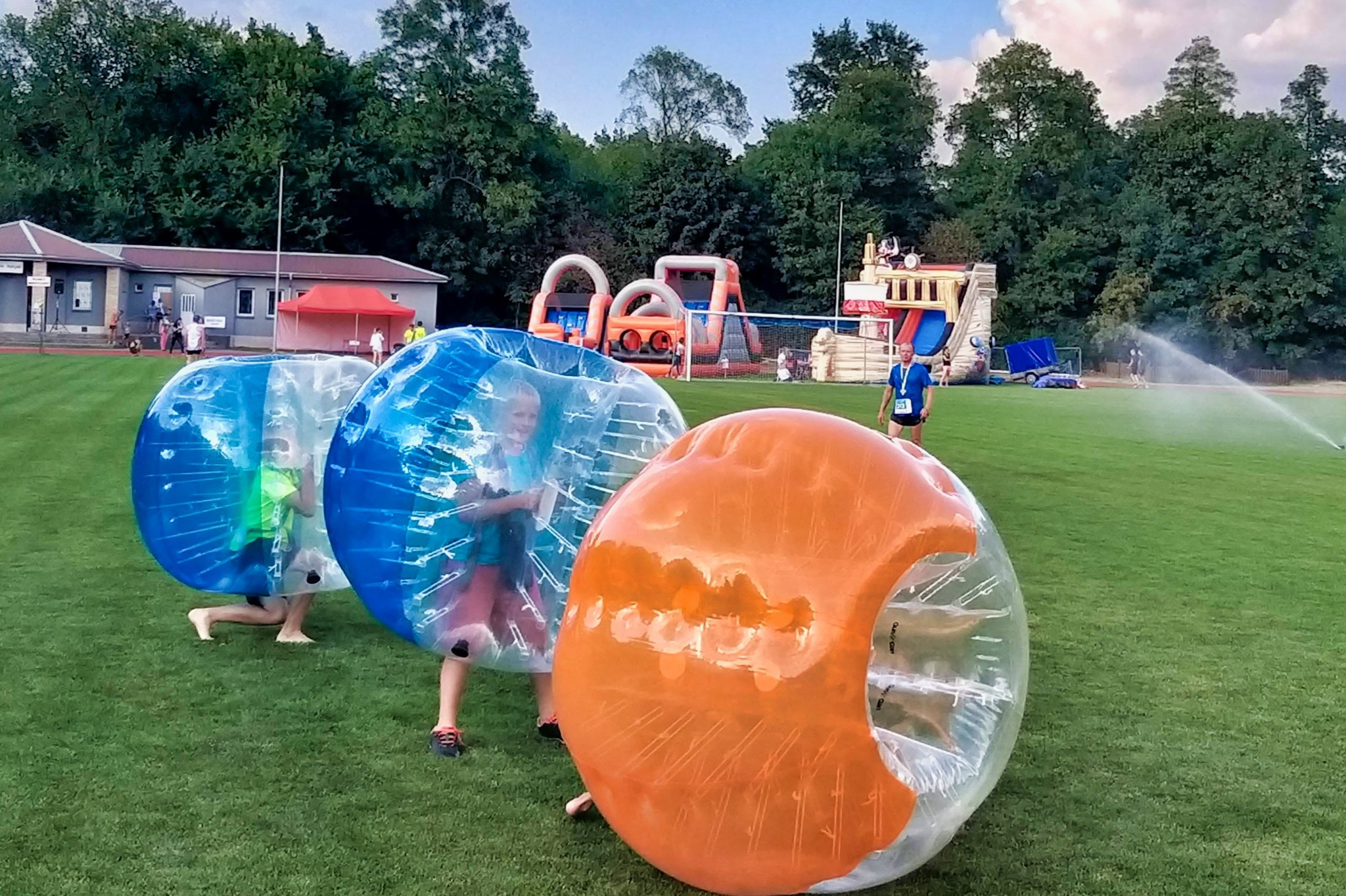 Dzieci w przezroczystych kulach bumper ball na zielonym boisku; w tle dmuchane zamki i zjeżdżalnie, a także budynek z czerwonym namiotem i zraszacz.