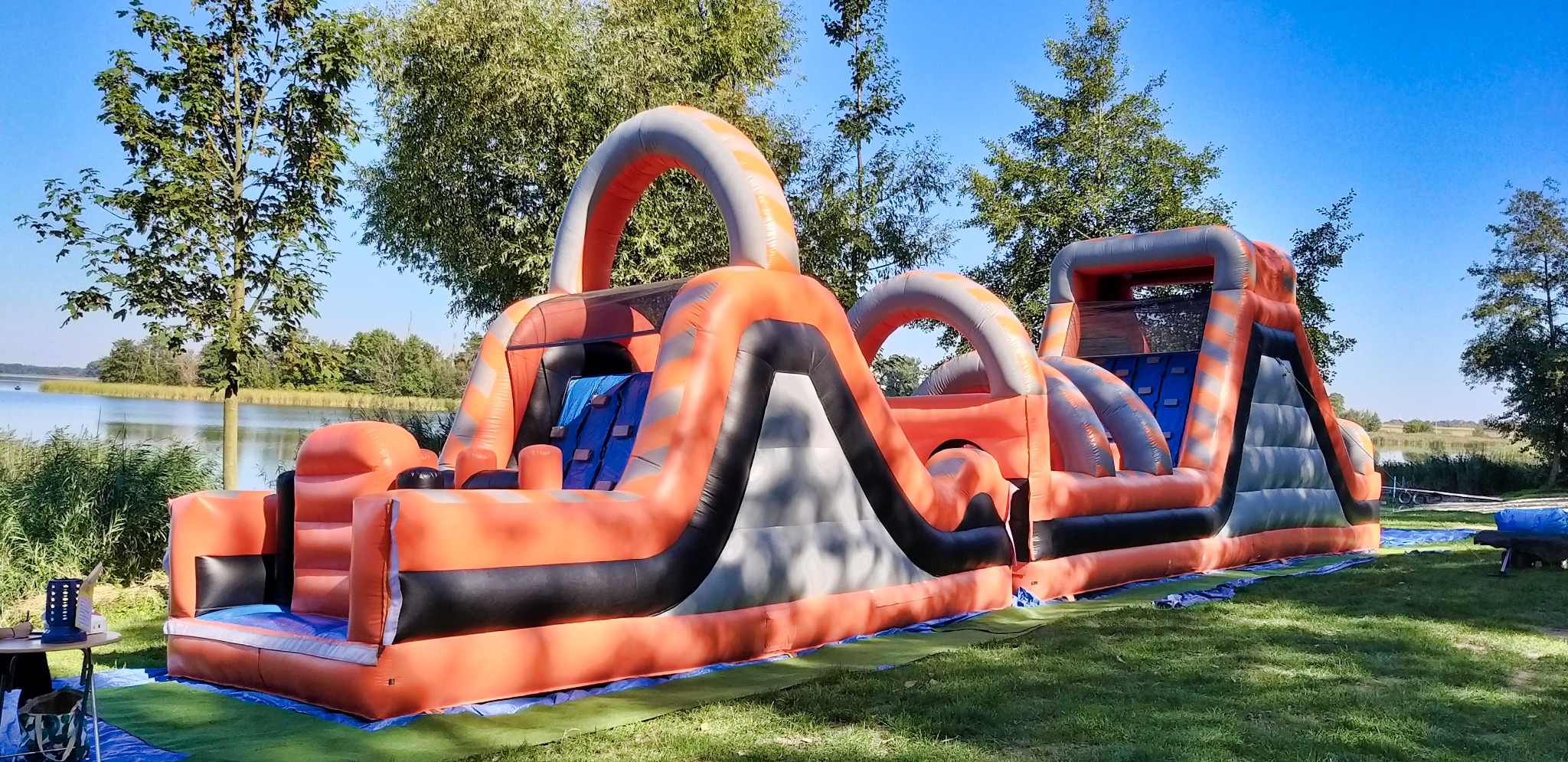 Duży, pomarańczowo-szaro-czarny dmuchany tor przeszkód z elementami wspinaczkowymi i zjeżdżalniami, rozstawiony na trawie w plenerze, z jeziorem i drzewami w tle, widoczny stół z akcesoriami po...