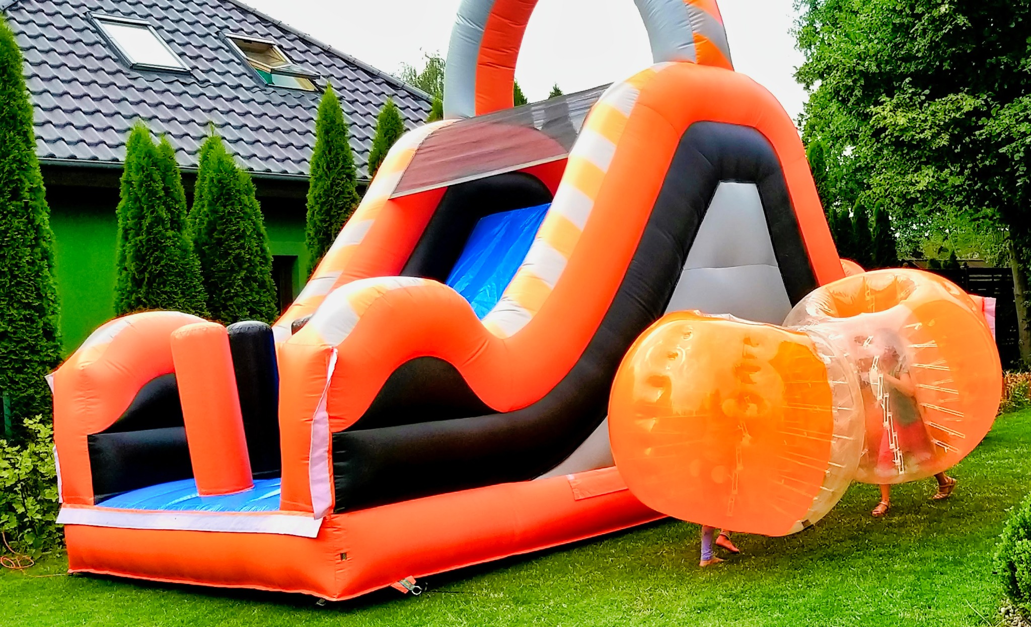 Pomarańczowo-czarny dmuchany zamek ze zjeżdżalnią, obok dwie osoby w pomarańczowych kulach sferycznych na trawniku przed zielonym żywopłotem i domem z szarym dachem.