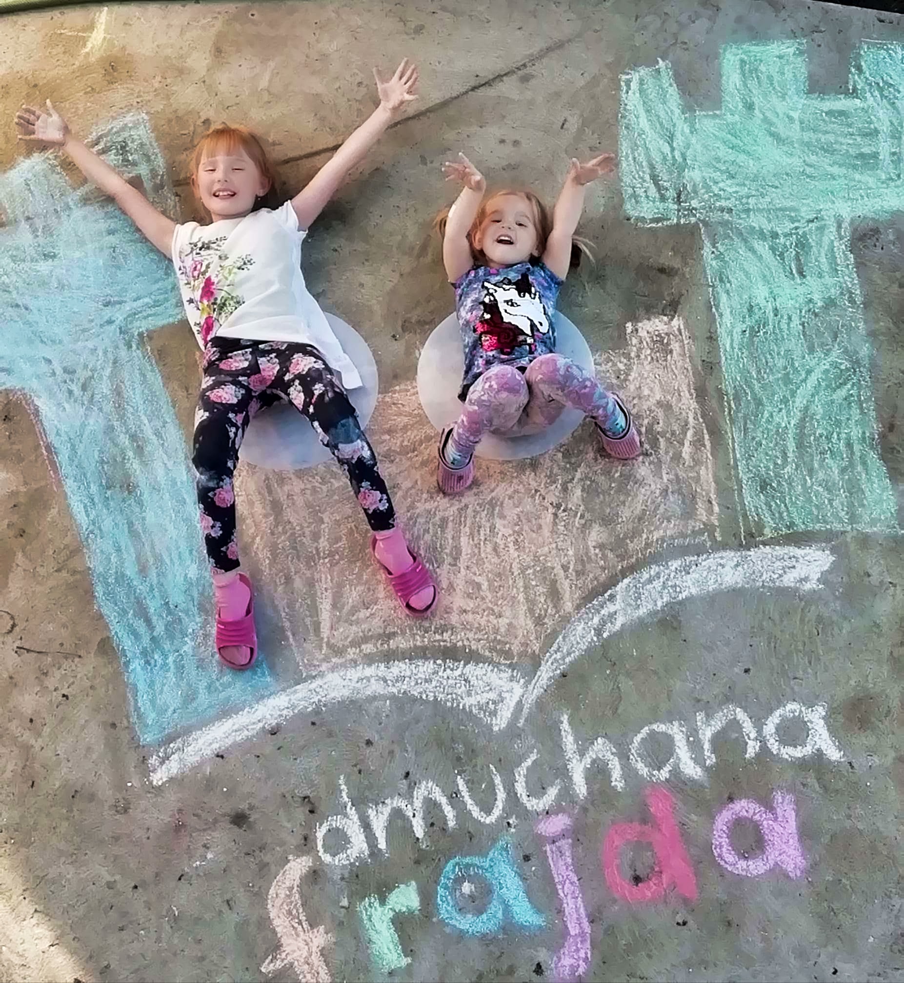 Dwie dziewczynki leżą na chodniku obok rysunków kredą: zamek i napis 'dmuchana frajda'.