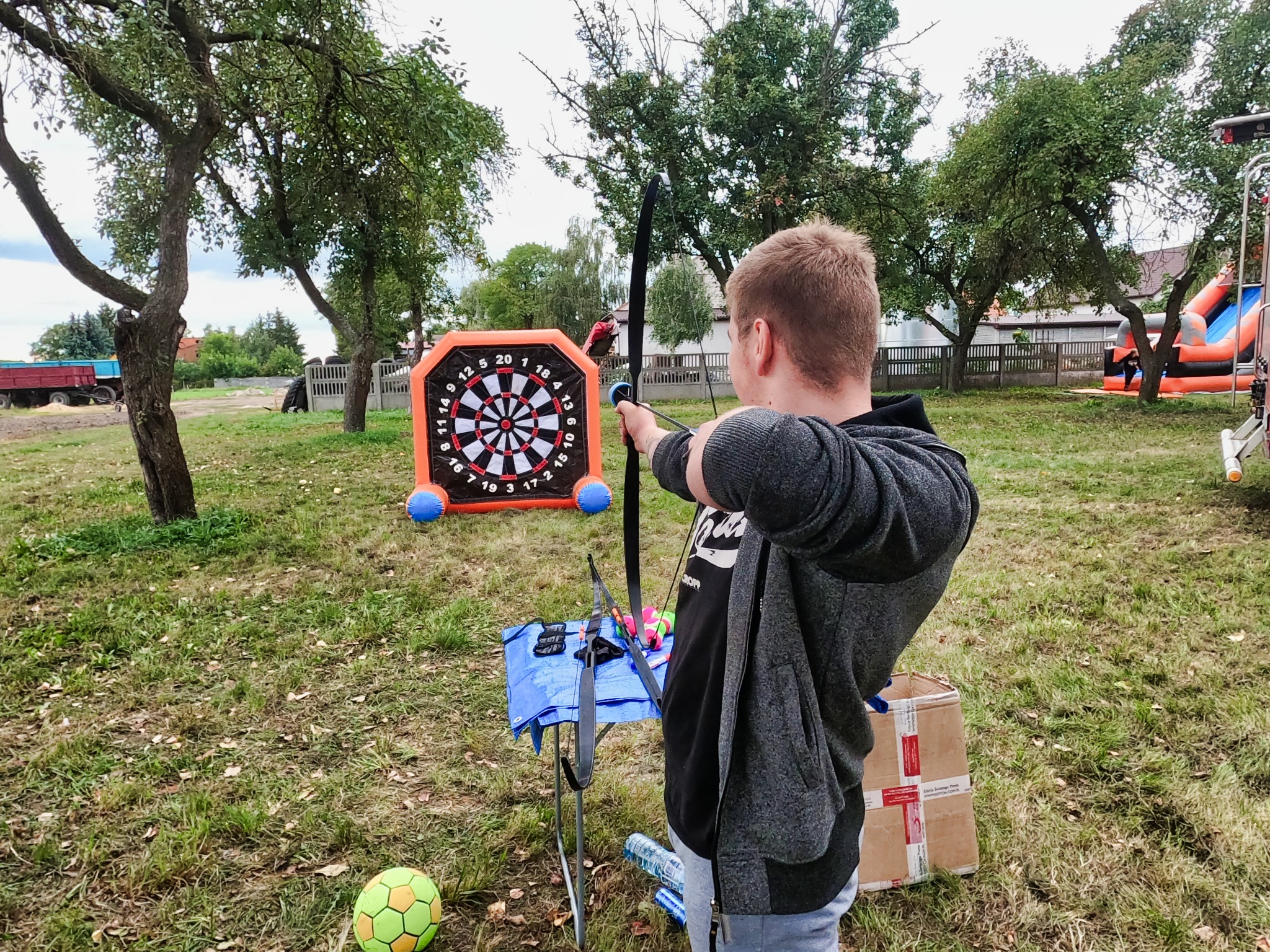 Chłopiec celuje z łuku do tarczy z dmuchaną obudową, widoczny dmuchaniec w tle, rozłożona arena do zabawy na trawie.