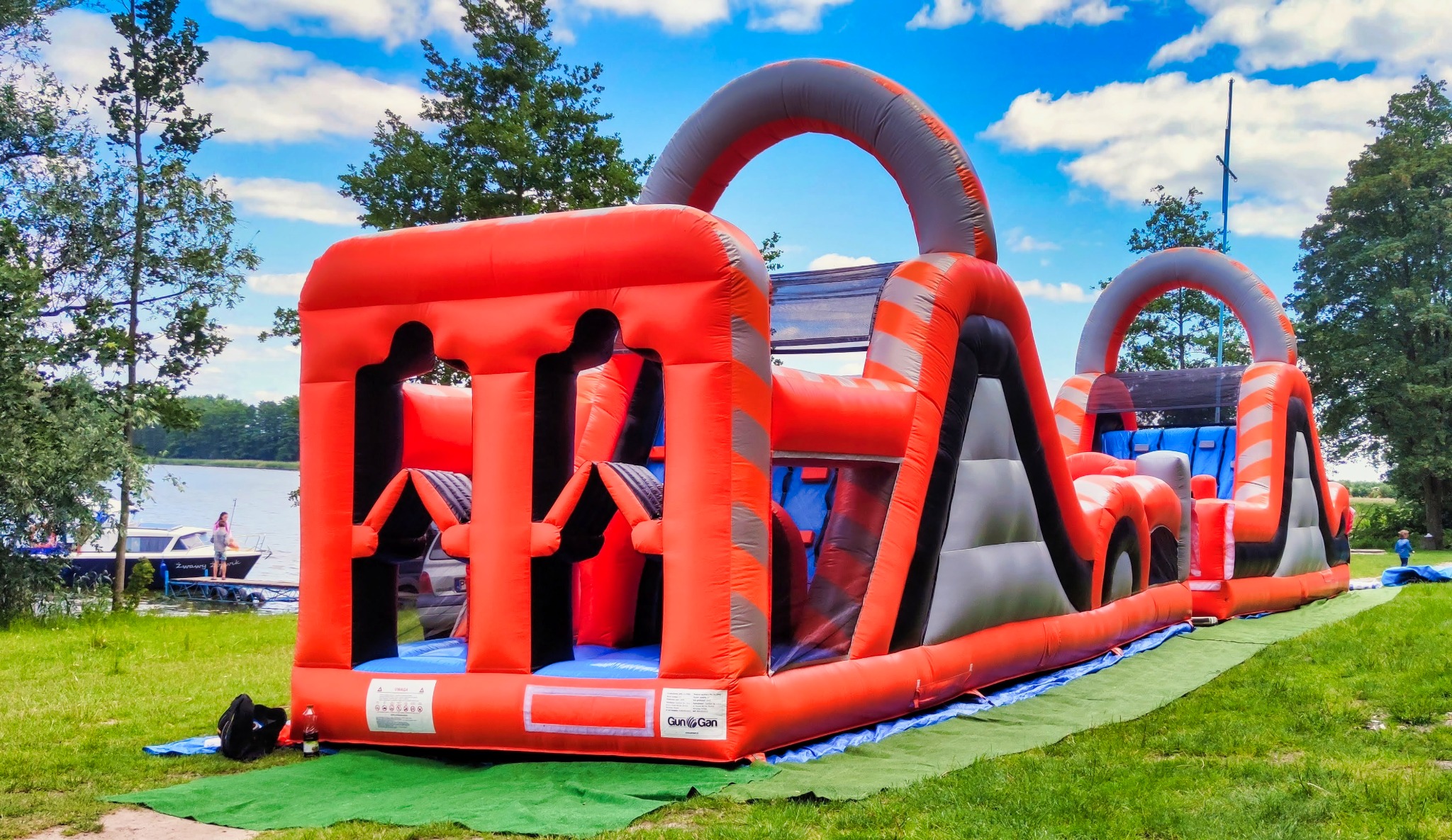 Rozłożony, dmuchany tor przeszkód w kolorach pomarańczowym, szarym i czarnym, ustawiony na trawie obok jeziora, z widocznym fragmentem łodzi i postacią w tle.