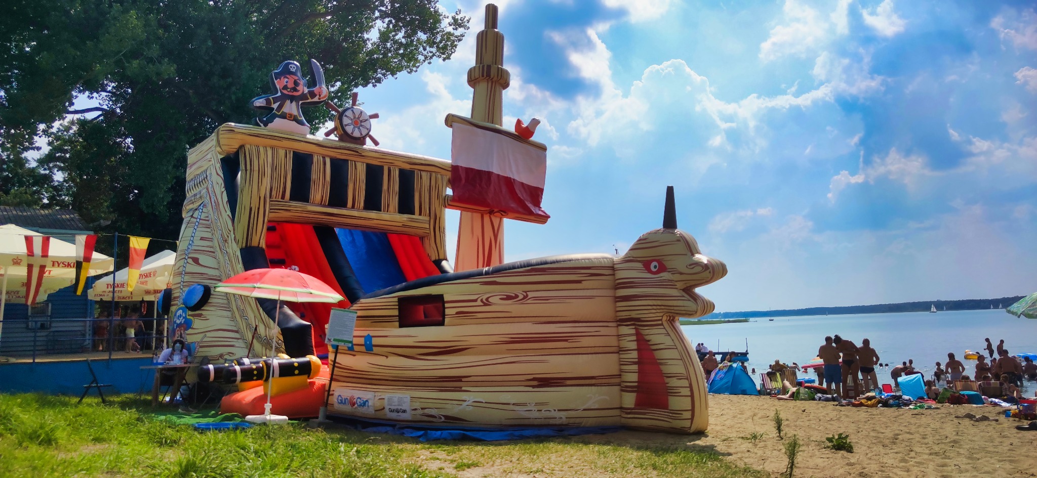 Nad jeziorem w Słupcy ustawiony dmuchany zamek w kształcie pirackiego statku z czerwoną zjeżdżalnią, na tle plażowiczów i błękitnego nieba z chmurami.