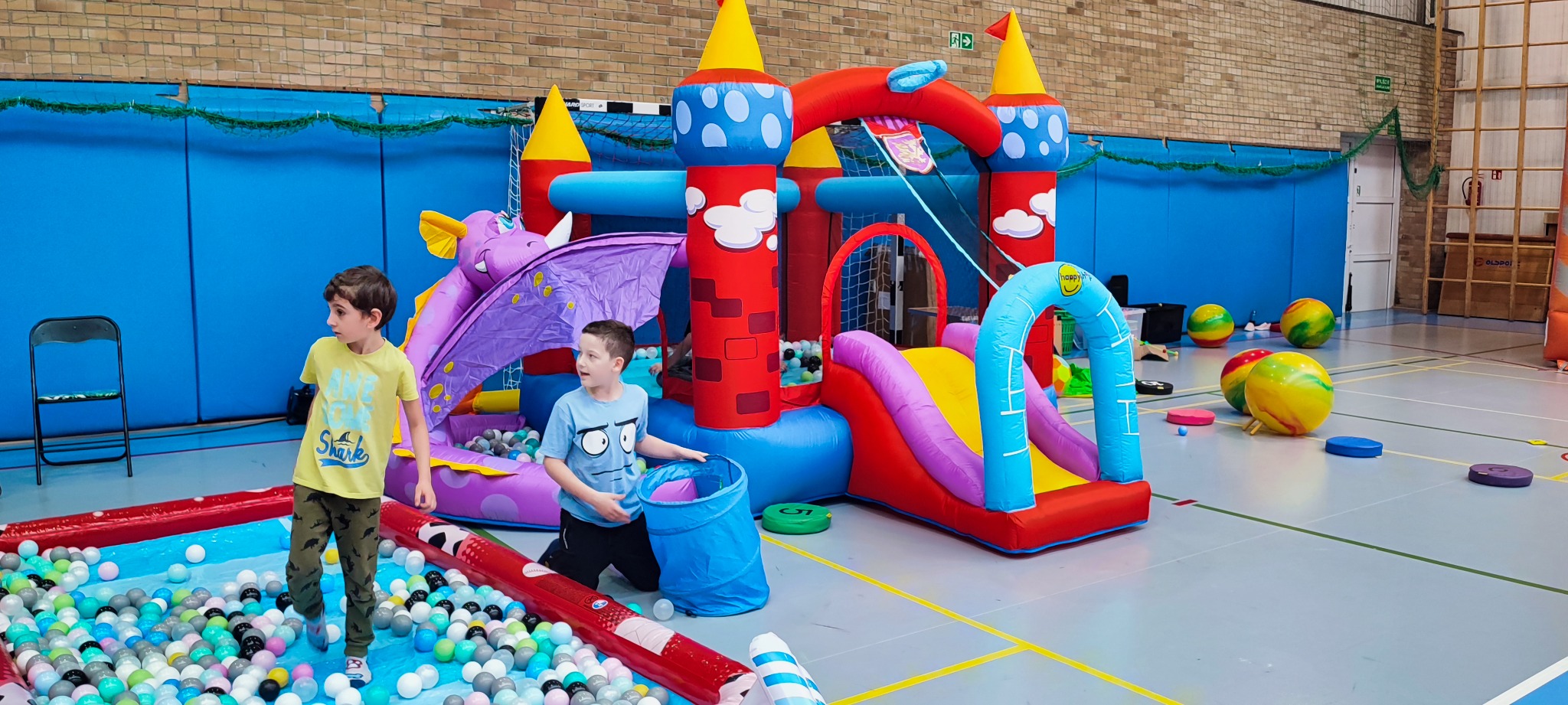Dwaj chłopcy bawią się w sali gimnastycznej przy dmuchanym zamku z motywem smoka, zjeżdżalnią i basenem z kulkami; jeden z nich wchodzi do basenu, drugi trzyma niebieski tunel.