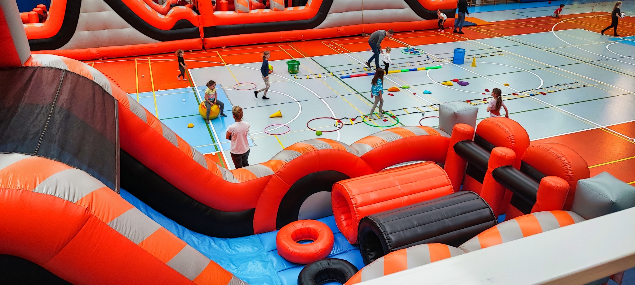 Wielki, pomarańczowo-szary dmuchany tor przeszkód w hali sportowej, dzieci pokonują różne przeszkody, w tle widoczny duży dmuchany zamek.