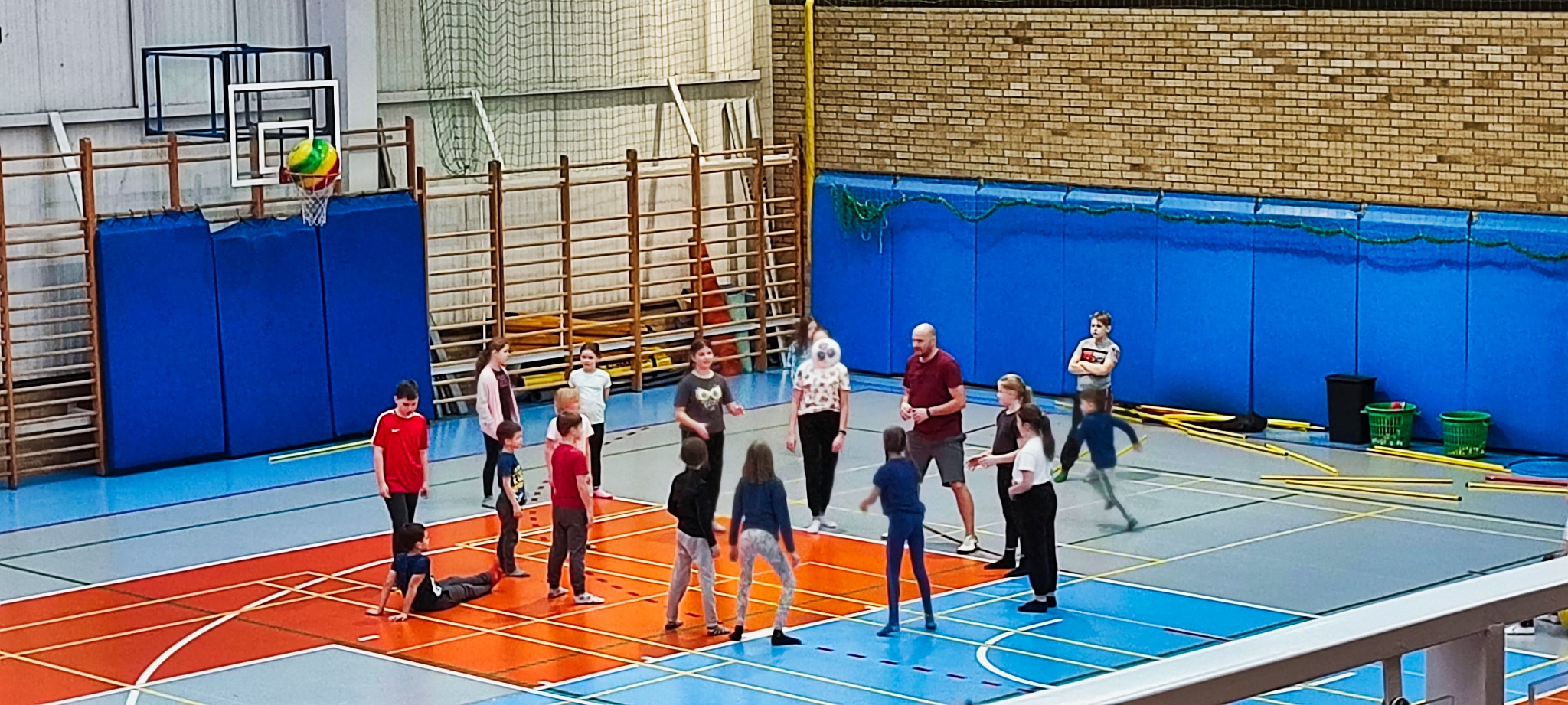 Grupa dzieci w różnym wieku i dorosły mężczyzna w hali sportowej podczas zajęć z elementami koszykówki i ćwiczeń zwinnościowych z użyciem tyczek.