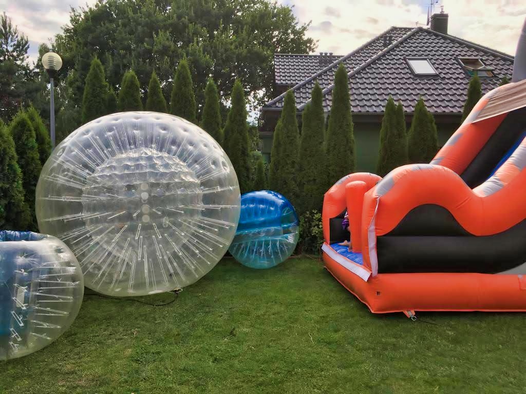 Pomarańczowy dmuchany zjeżdżalnia i przezroczyste kule zorbingowe na trawie przed domem z ciemnym dachem i tujami w tle.