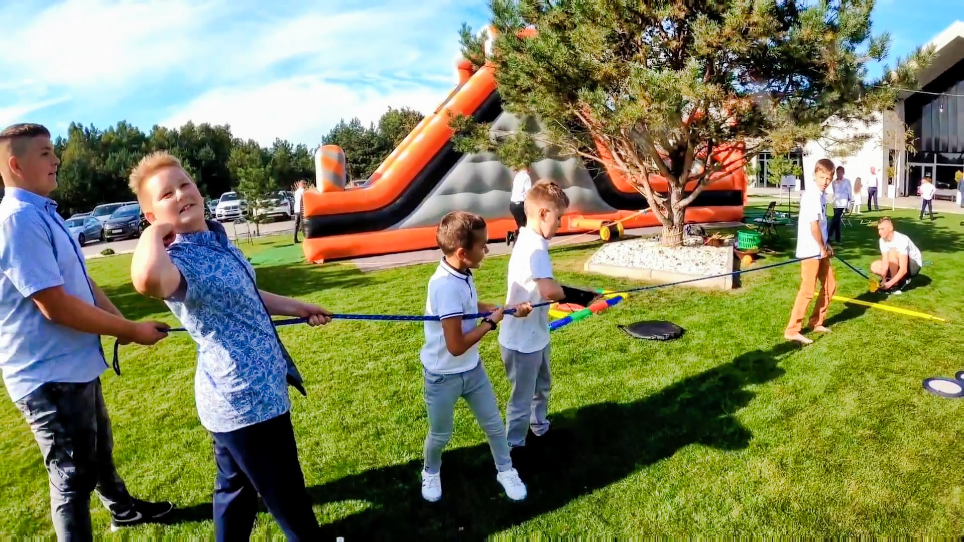 Grupa dzieci gra w przeciąganie liny na zielonym trawniku, w tle pompowana, pomarańczowo-szara zjeżdżalnia i budynek.