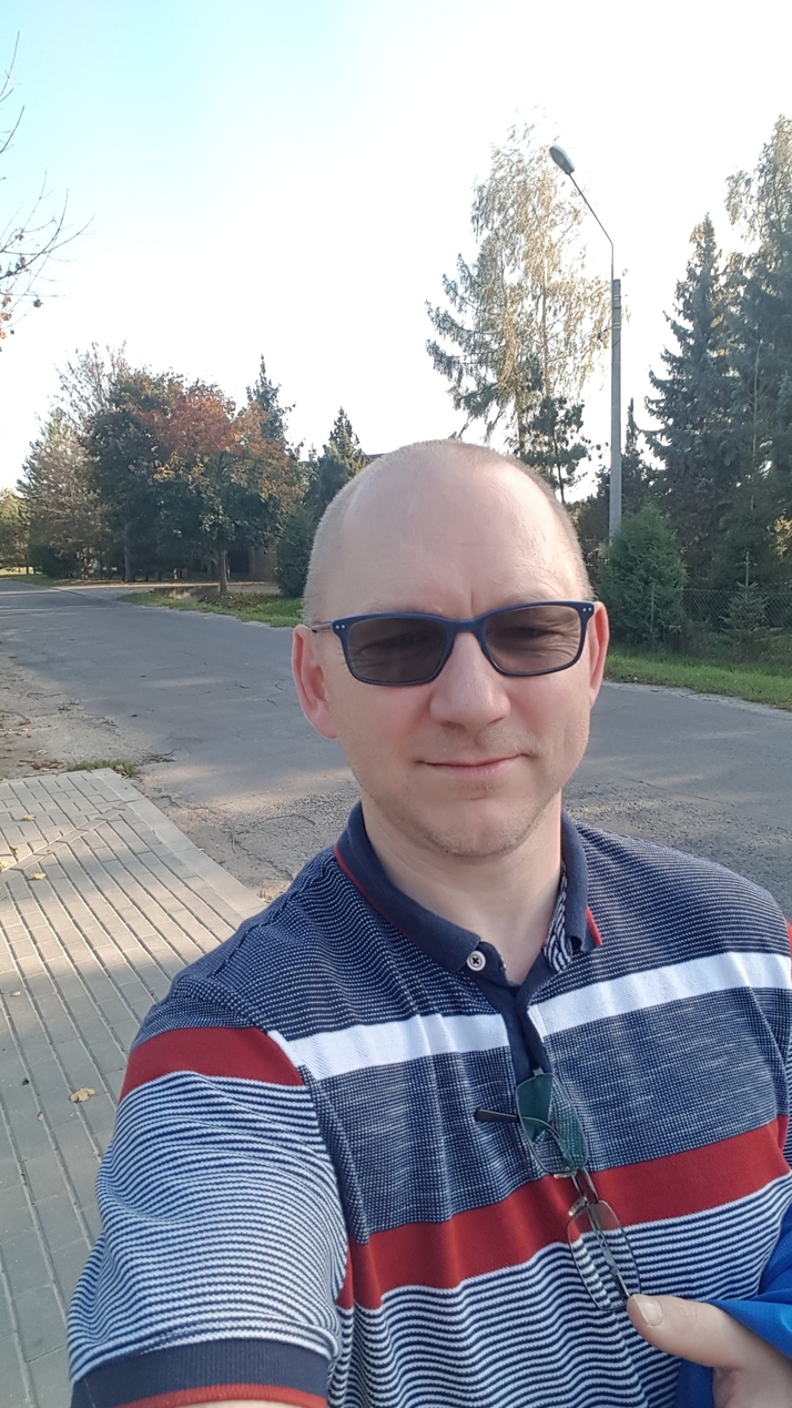 Mężczyzna w okularach przeciwsłonecznych, na zewnątrz, w tle ulica i drzewa. Autoportret.