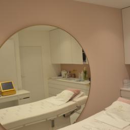 MJO Profibuild - Refleksja w okrągłym lustrze ukazuje wnętrze gabinetu kosmetycznego z dwoma białymi leżankami, jasnoróżowymi ścianami i białymi szafkami. Na jednej z leżanek widoczny ręcznik z logo.