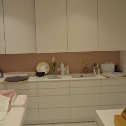 MJO Profibuild - Jasne wnętrze gabinetu kosmetycznego z białymi szafkami, różowym akcentem na ścianie i złotymi detalami, takimi jak lustro i armatura, z leżanką kosmetyczną z ręcznikiem z logo i sprzętem...