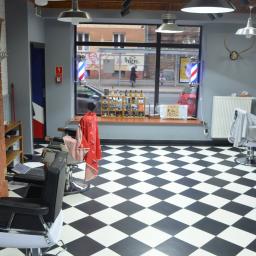 MJO Profibuild - Wnętrze zakładu fryzjerskiego z czarno-białą szachownicą na podłodze, widoczne fotele fryzjerskie, dekoracje ścienne w postaci poroża oraz okno z widokiem na ulicę z charakterystycznymi słupkami...