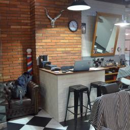 MJO Profibuild - Wnętrze nowoczesnego salonu barberskiego z recepcją wykończoną białymi płytkami, brązową cegłą na ścianie, skórzaną sofą i czarno-białą szachownicą na podłodze, widoczne stanowiska fryzjerskie...