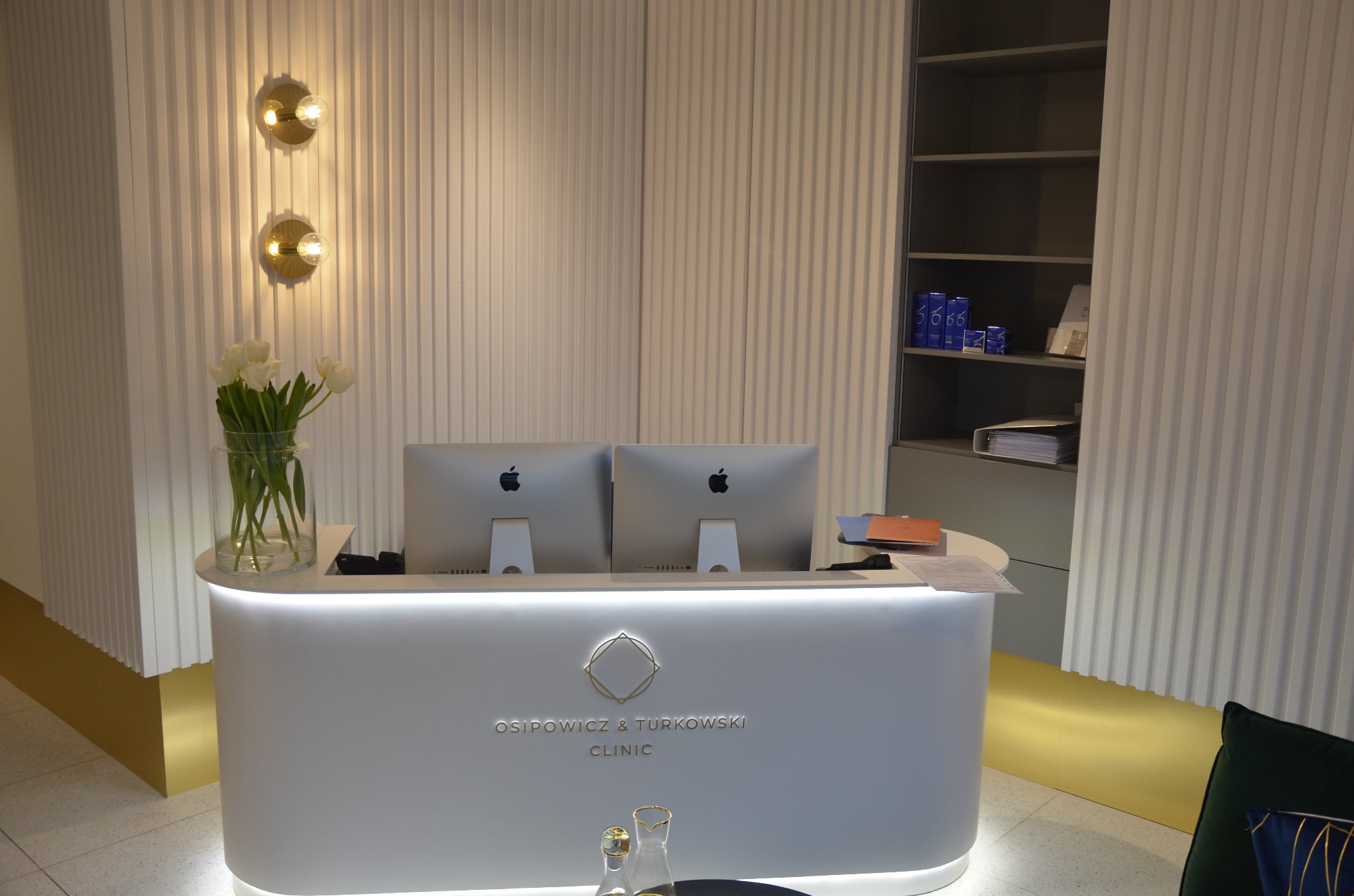 Recepcja kliniki z białym, podświetlanym biurkiem, dwoma monitorami Apple, wazonem z białymi tulipanami i oświetleniem ściennym w formie złotych kul na tle ściany z pionowymi panelami.