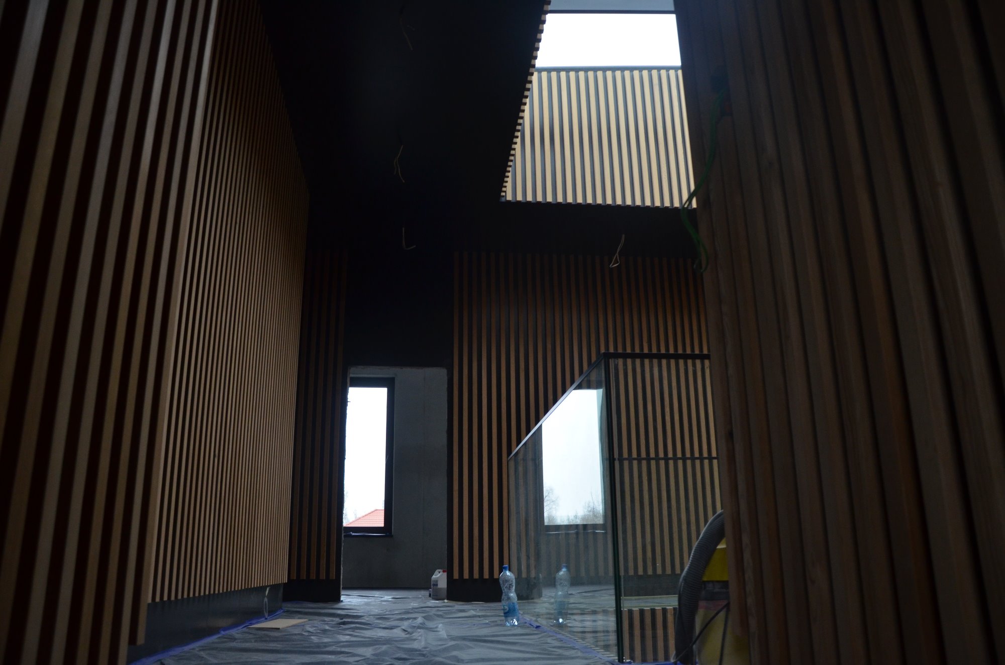 Wnętrze w trakcie prac wykończeniowych: pionowe panele drewniane na ścianach i suficie, zabezpieczona folią podłoga, szklana balustrada schodów, okno z widokiem na zewnątrz.