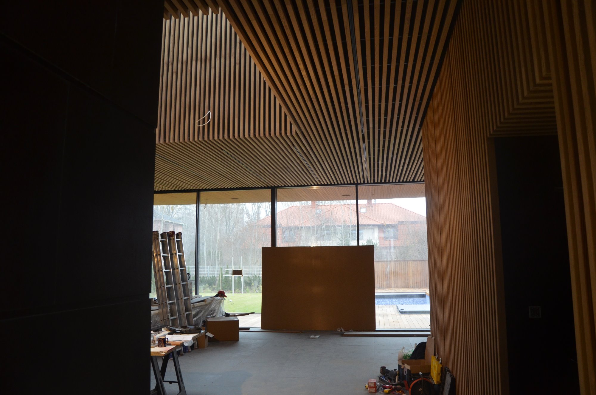 Wnętrze w trakcie prac wykończeniowych: drewniane lamele na suficie i ścianie, duże okno z widokiem na ogród z basenem, drabina oparta o ścianę, narzędzia na podłodze.