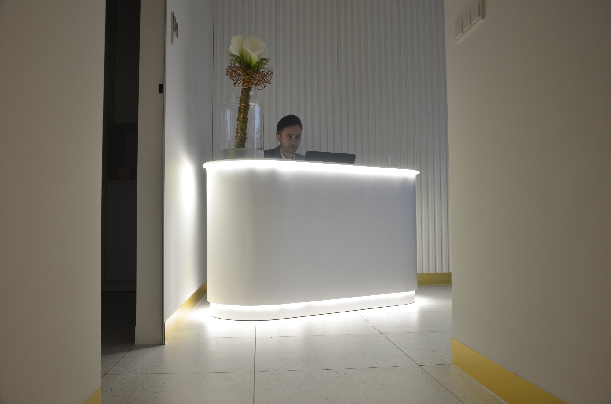 Recepcja biurowa z podświetlanym, białym blatem, za którym siedzi mężczyzna, na blacie wazon z białą kalą, ściana z pionowymi żłobieniami.
