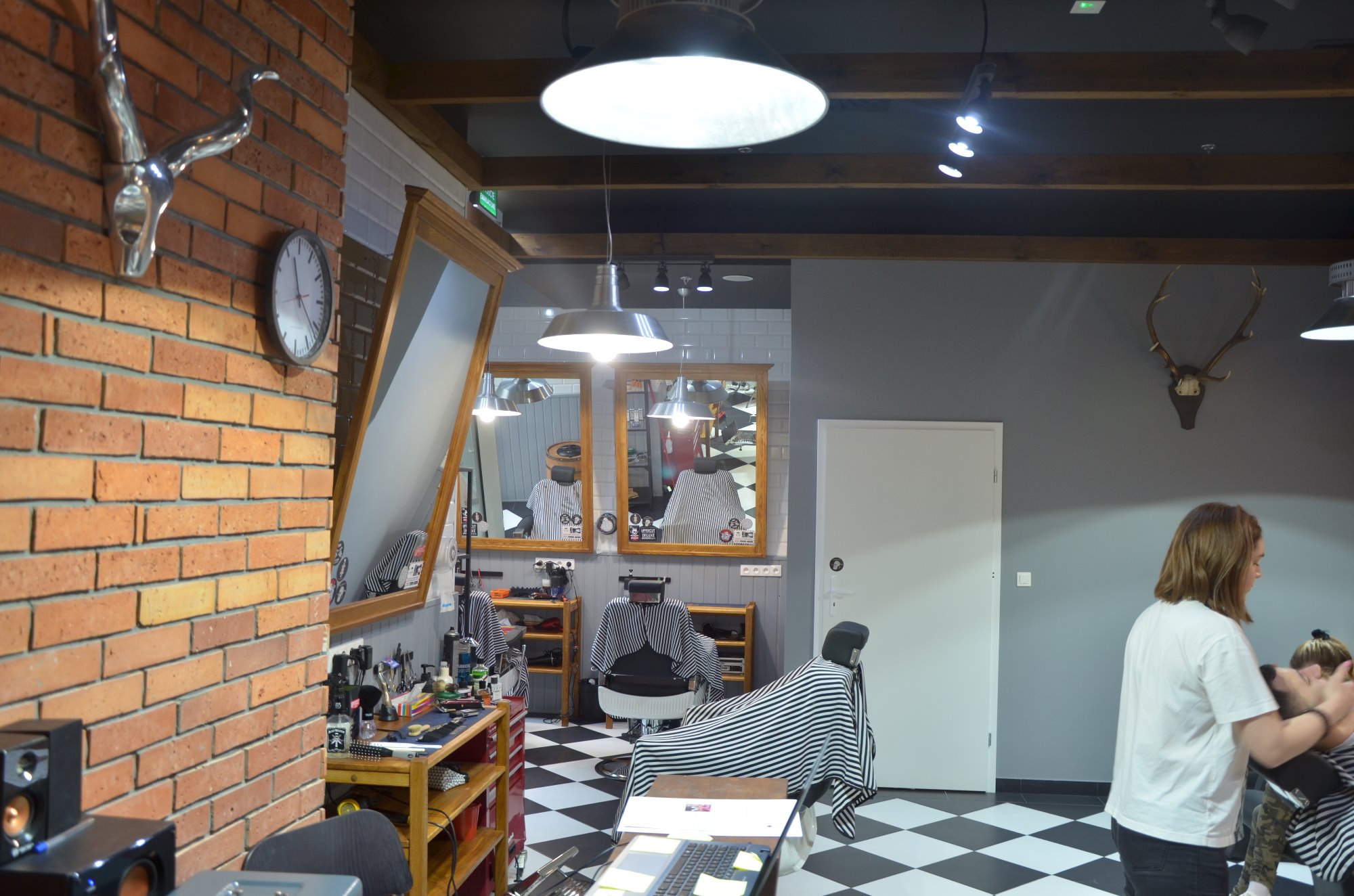 Wnętrze nowoczesnego salonu fryzjerskiego z ceglaną ścianą, dekoracjami w postaci głów zwierząt, dużymi lustrami w drewnianych ramach, czarno-białą podłogą w szachownicę i stanowiskami pracy...