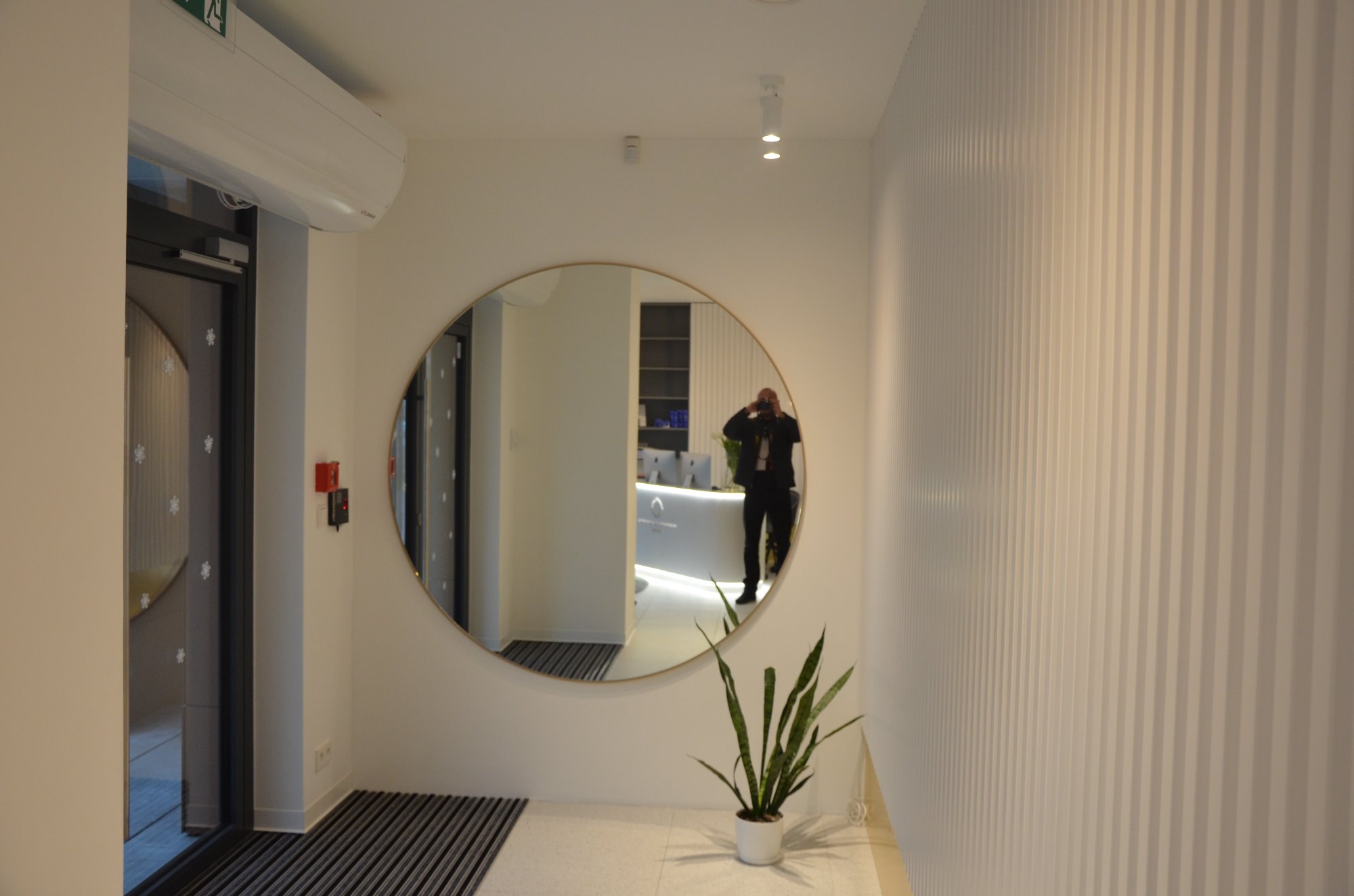Nowoczesne wejście do biura z okrągłym lustrem odbijającym recepcję i fotografa, minimalistyczna dekoracja ścian z pionowymi paskami, roślina doniczkowa w białej osłonce.