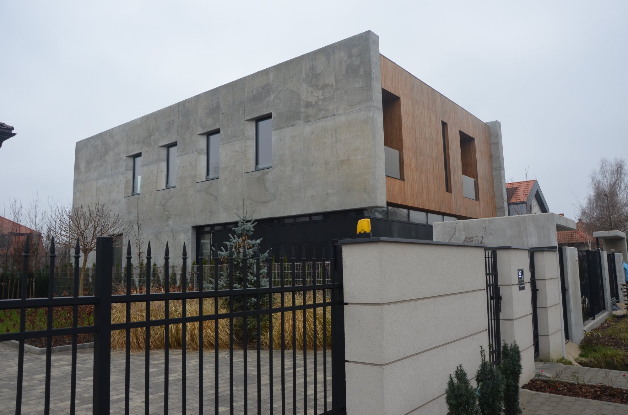 Nowoczesny dom jednorodzinny z betonu architektonicznego i drewnianą elewacją, widoczny zza czarnego, kutego ogrodzenia i betonowego murku z numerem domu.