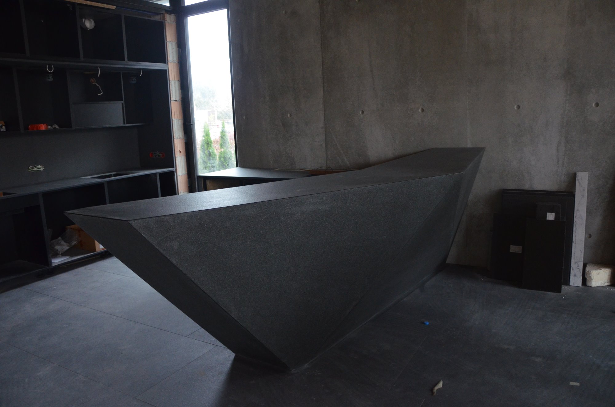 Surowa, nowoczesna kuchnia w trakcie wykończenia: monolityczna, grafitowa wyspa kuchenna o geometrycznym kształcie, na tle betonowej ściany i czarnych szafek kuchennych bez frontów, widoczne...